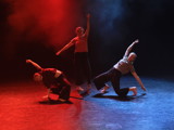 Bildet viser tre dansere på scene. De står i en trekant. Hun bakerste strekker seg opp, mens de to framderste er på vei i ett fall ned til gulvet. Lyset er blått og rødt og det er litt røyk på scenen. 