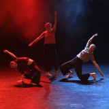 [Bildet viser tre dansere på scene. De står i en trekant. Hun bakerste strekker seg opp, mens de to framderste er på vei i ett fall ned til gulvet. Lyset er blått og rødt og det er litt røyk på scenen.] 