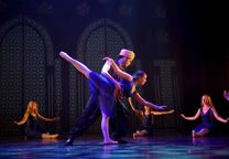 Bildet viser to dansere i en duett på scenen. De er prins og prinsesse. Hun bøyer seg framover med beinet høyt opp bak. Kjolen er dyp blå. 