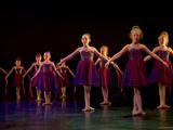 Bildet viser ni ballettdansere på scene. De har lilla kjole på og står i 3. posisjon med arner over skjørtene. 